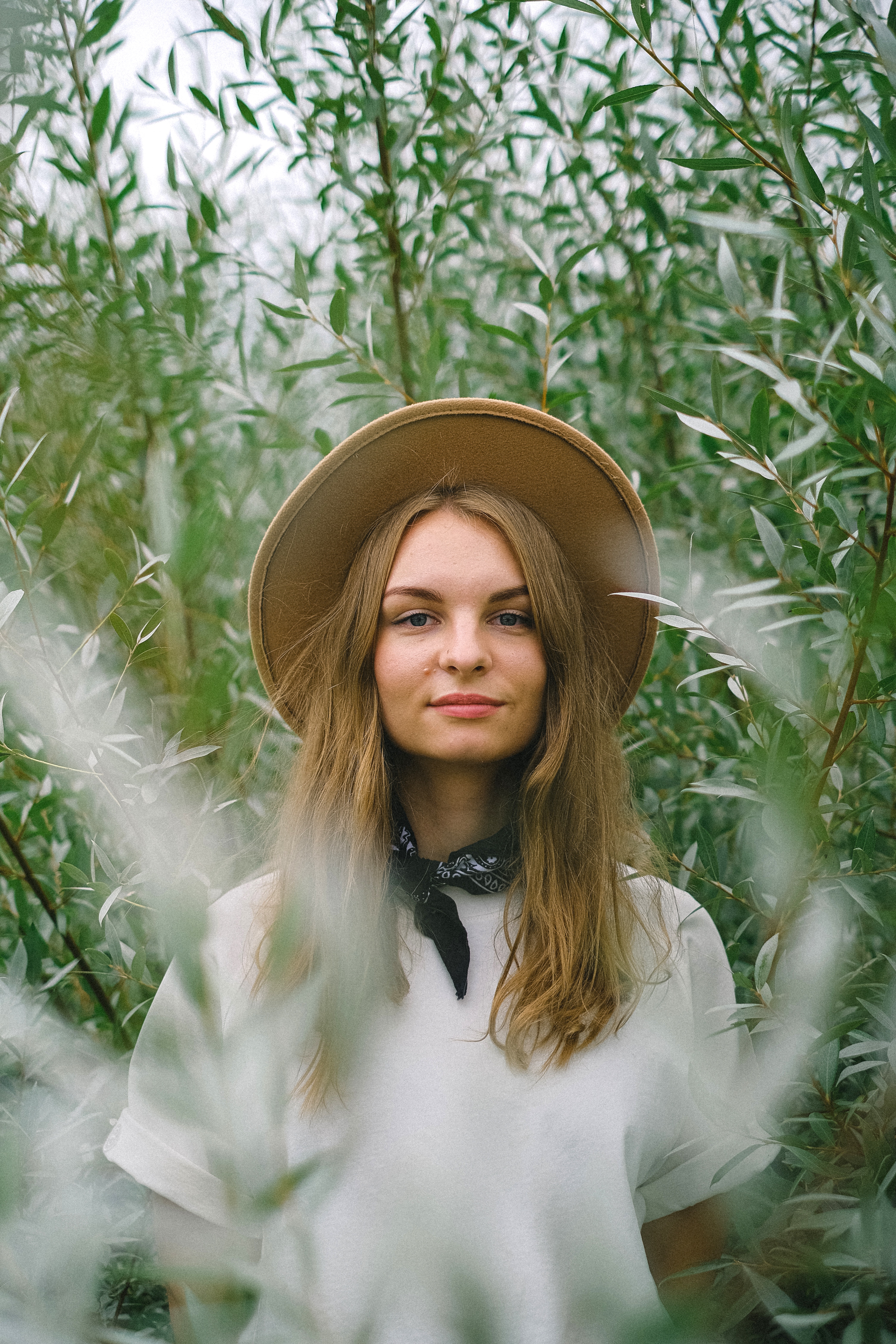 a woman staring at camera with greenery behind her and is wearing a hat